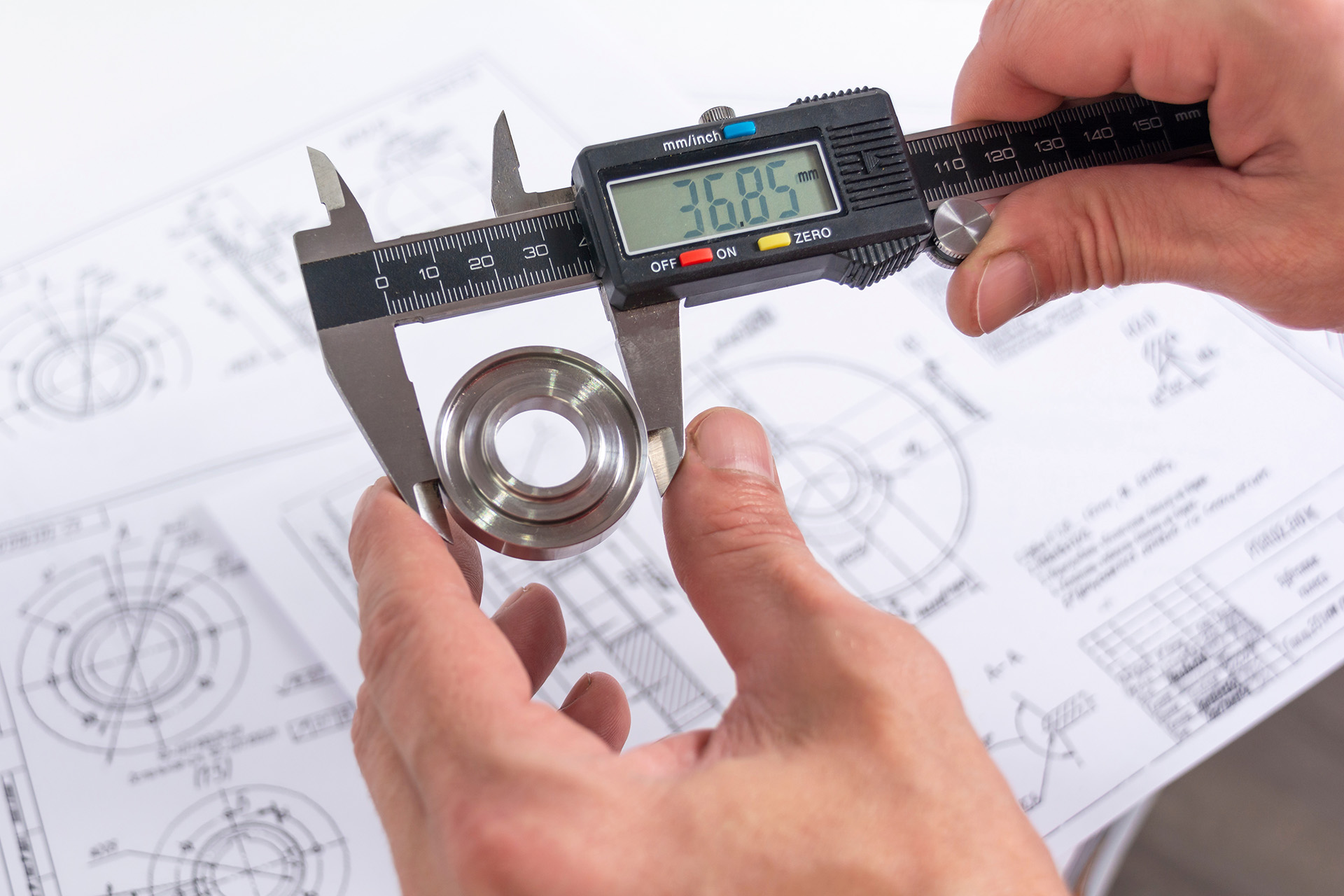 Hands of an engineer measures a metal part with a digital vernier caliper against the background of technical drawings. Quality control of parts machined on a lathe. Hands of an engineer measures a metal part with a digital vernier caliper against the background of technical drawings. Quality control of parts machined on a lathe.